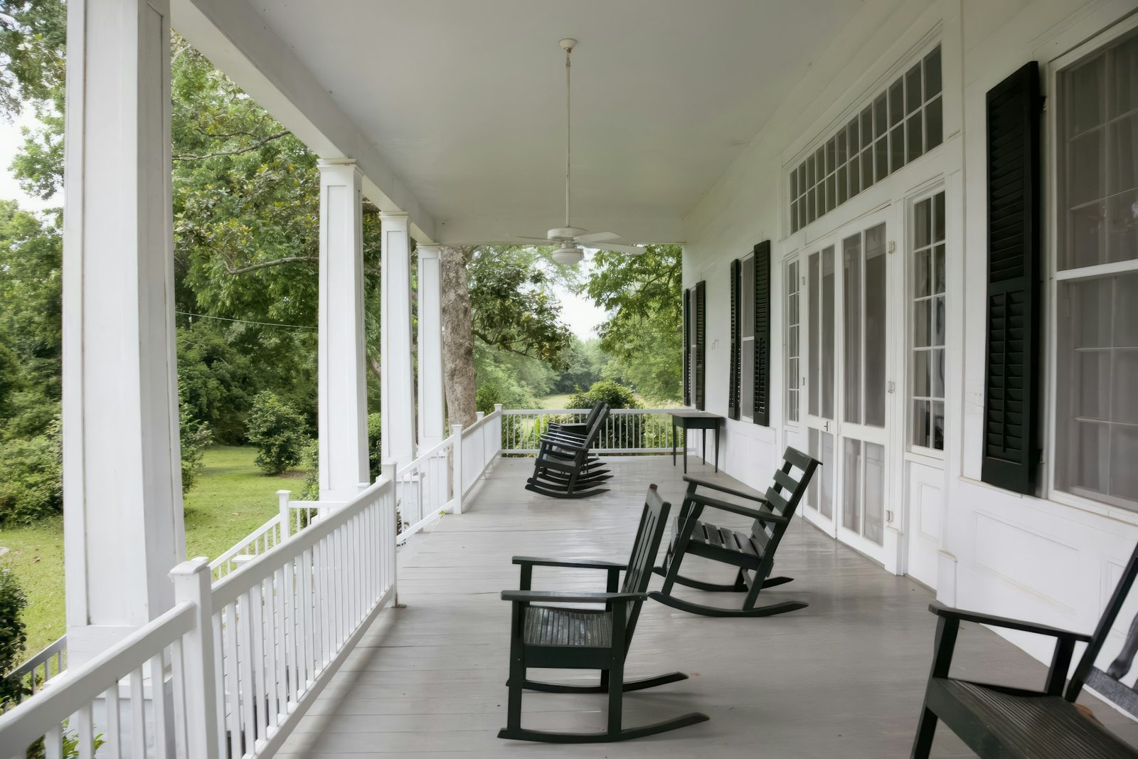 Deep covered porch of a white Southern house, with green rocking chairs, square columns and shuttered doors opening onto a shaded lawn